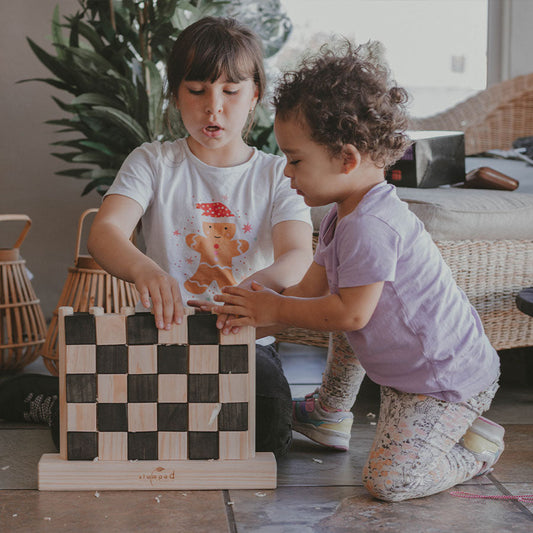 Two children playing with a wooden checkered toy in a home setting.