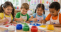 Four children painting at a table with colorful paint and brushes in a classroom setting.